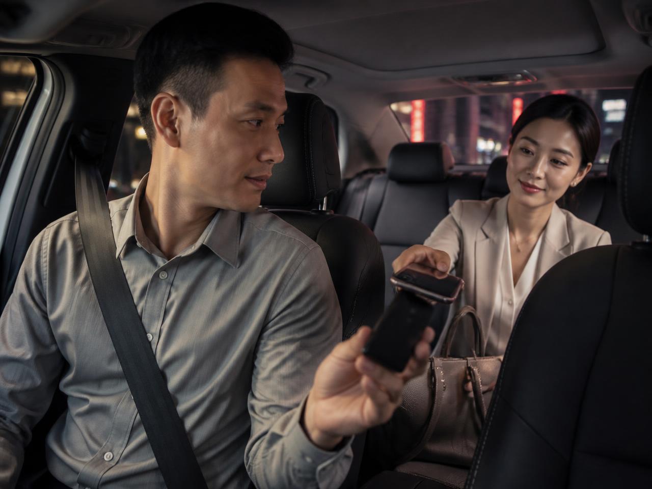 Asian private hire driver holding his smartphone toward the back seat while a passenger taps her phone against it to pay at the end of the trip
