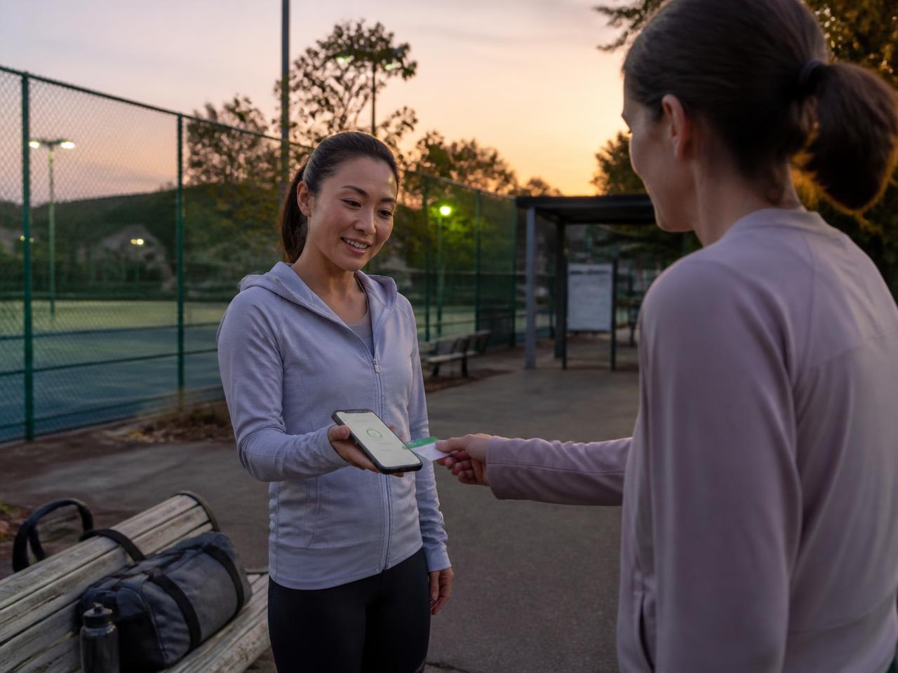 Asian fitness coach at an outdoor sports court in Japan holding out her smartphone while her client taps a contactless card on it after the session